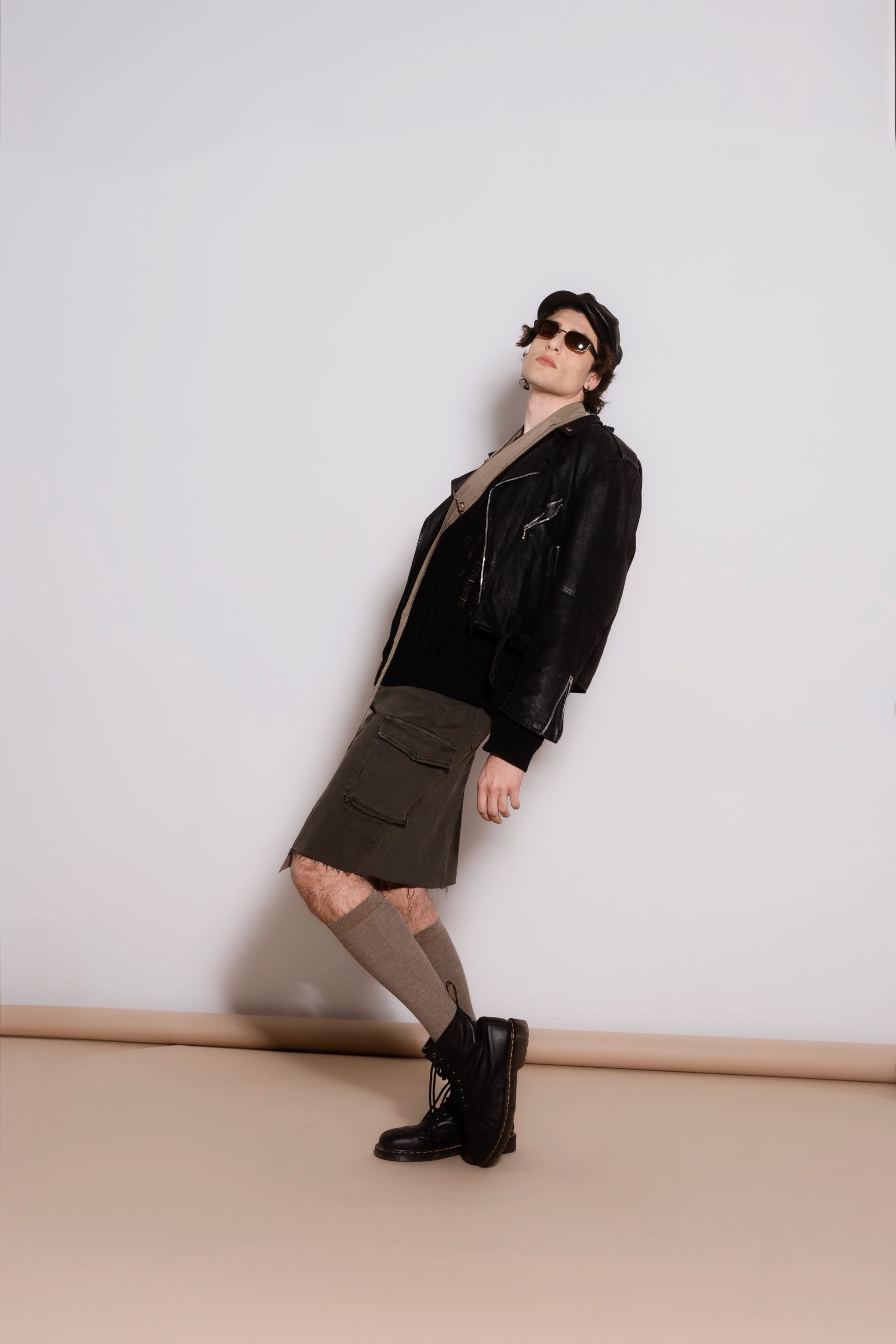 Modello maschile in giacca di pelle nera e gonna cargo verde, abbinato a calze e stivali, stile urbano innovativo.