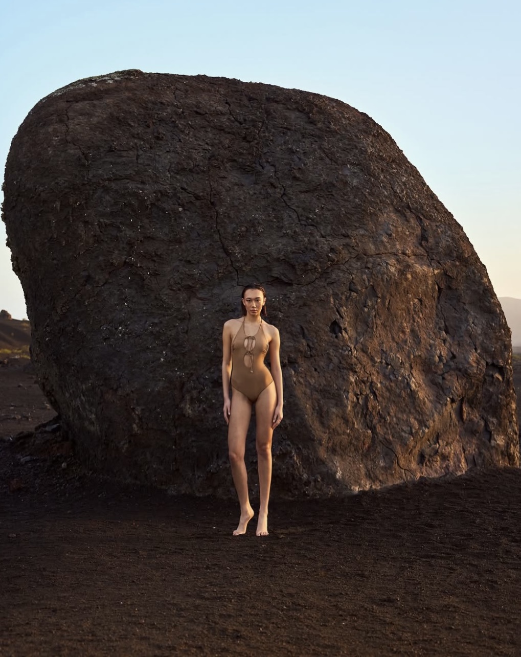 Modella in costume adamitico davanti a una grande roccia in un paesaggio vulcanico, stile fotografico contemporaneo.
