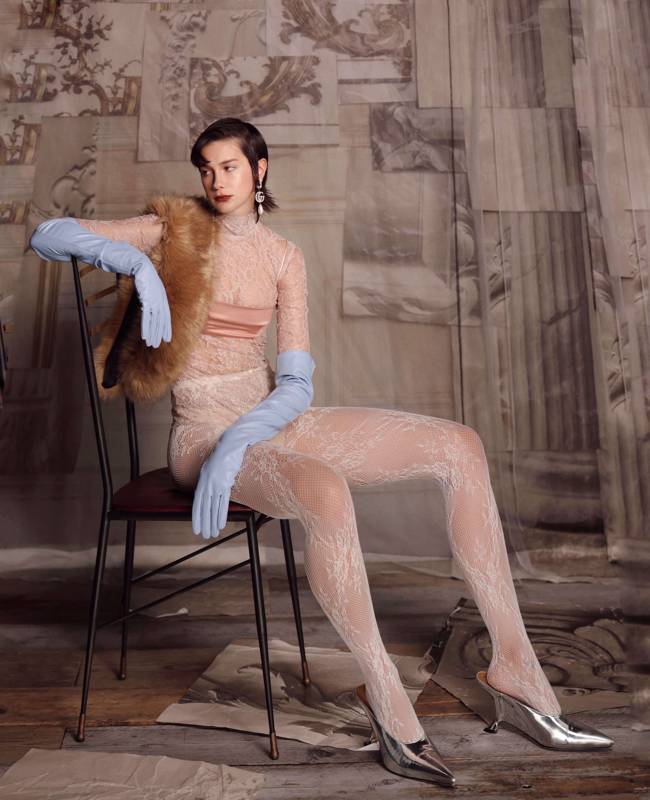 Modella elegante in abito in pizzo, guanti blu e scarpe argentate, posa su sedia con sfondo artistico.