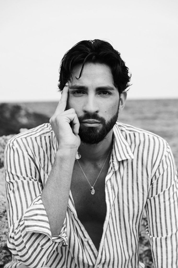 Modello maschile in camicia a righe, sguardo intenso sullo sfondo marino, fotografia in bianco e nero.