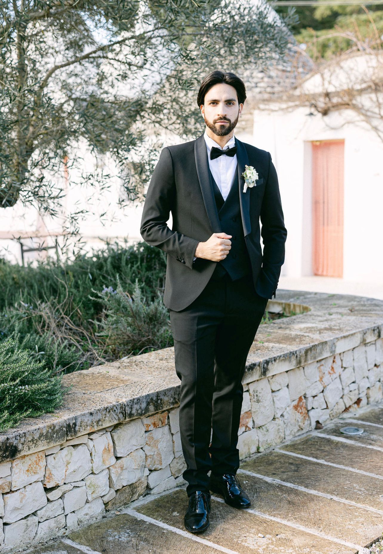 Uomo elegante in abito da cerimonia con cravatta nera e boutonnière, posizionato in un'ambientazione naturale.