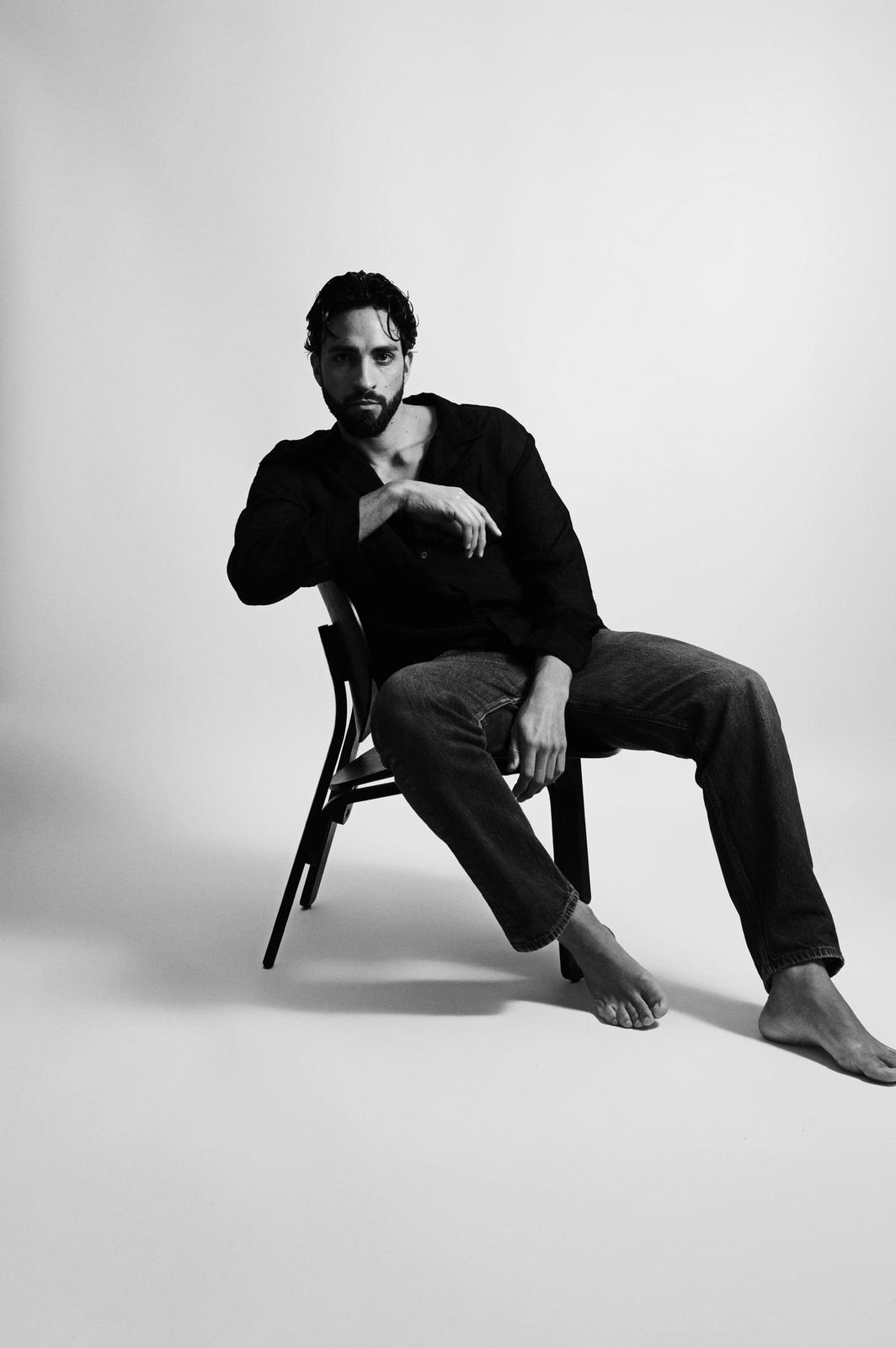 Modello maschile seduto su una sedia nera, sguardo intenso, stile elegante e casual, fotografo di moda.