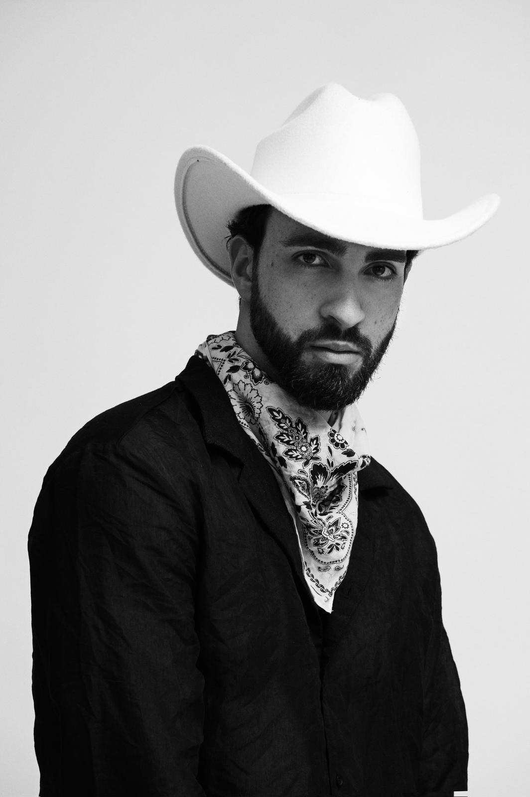 Modello in look cowboy con cappello bianco e foulard, stile maschile raffinato e audace per servizi fotografici di moda.