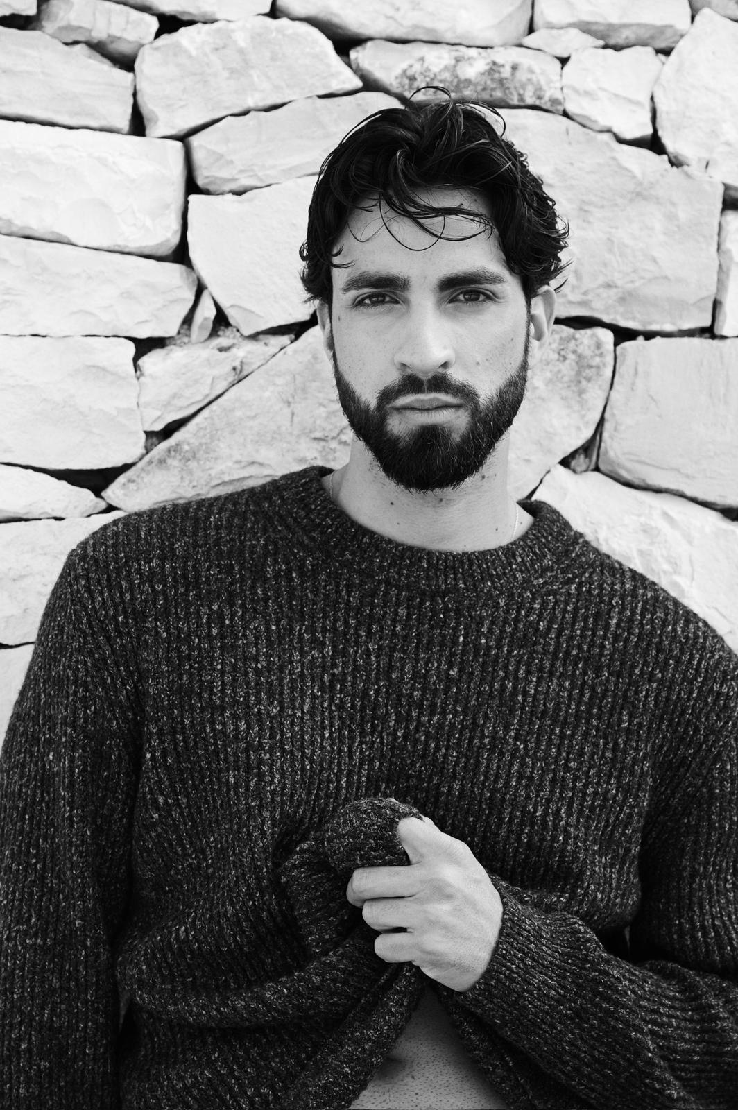 Modello maschile con maglione grigio scuro davanti a una parete di pietra, espressione intensa e sofisticata.
