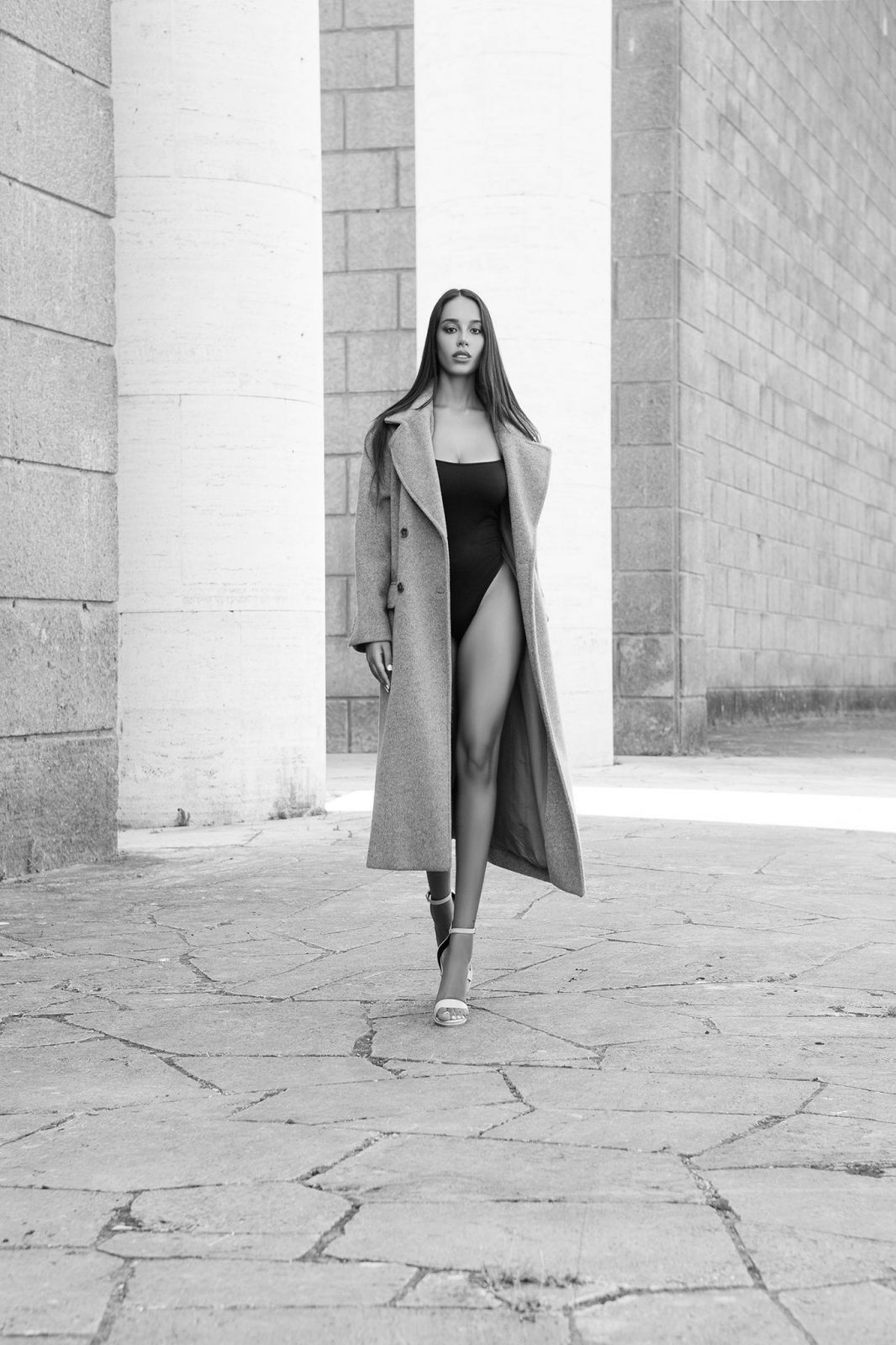 Modella elegante con cappotto grigio e costume, cammina tra colonne in ambientazione urbana, stile fashion raffinato.