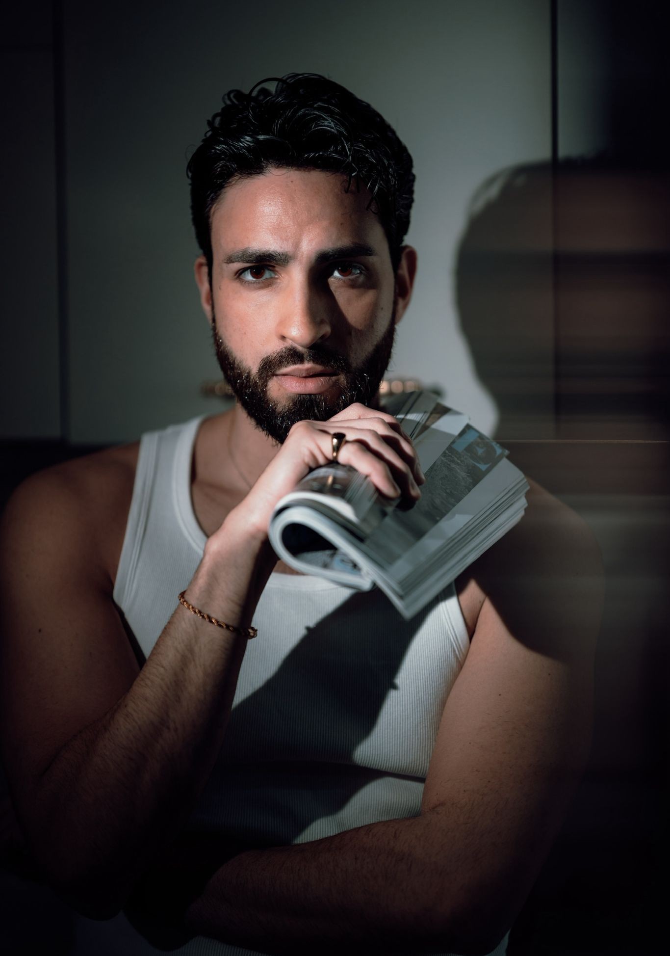 Uomo affascinante con barba, in t-shirt bianca, tiene una rivista di moda in un'atmosfera elegante e misteriosa.